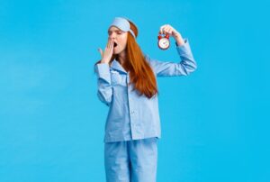 Sleepy woman in pajamas holding an alarm clock and yawning | Featured image for the "Stages of Sleep Part 2" blog post.