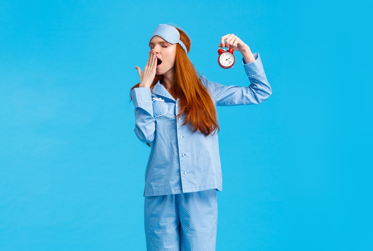 Sleepy woman in pajamas holding an alarm clock and yawning | Featured image for the "Stages of Sleep Part 2" blog post.