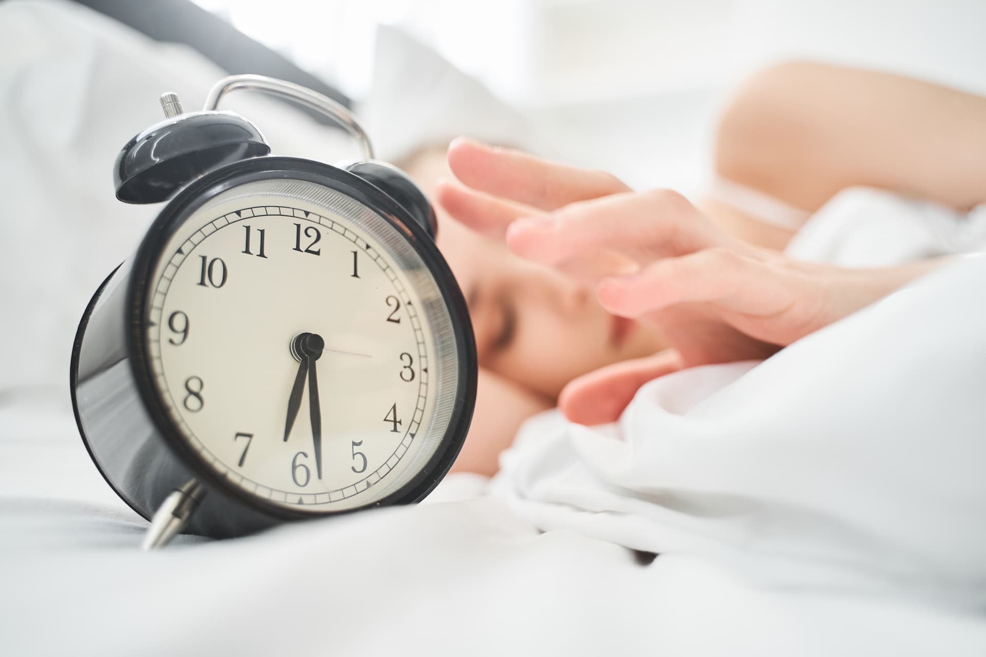 A young woman resting in bed next to an alarm clock| Featured image for the "How Lack of Sleep Affects the Body" blog post.