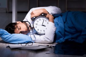 Man sleeping and holding a giant alarm clock | Featured image for the Shift Work Sleep Disorder – Sleeping Against the Clock Wenatex blog from Wenatex.